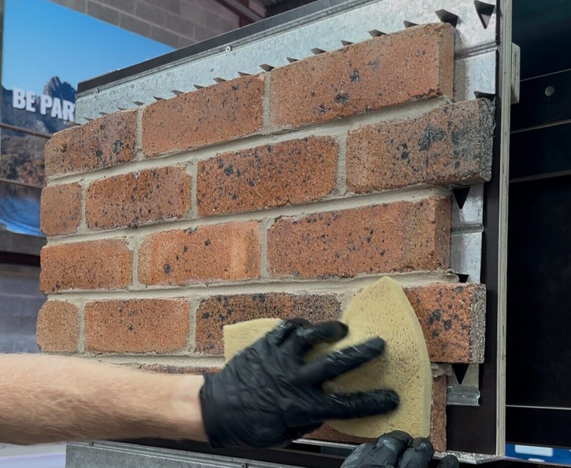 A person wearing black gloves uses a yellow sponge to clean or smooth recycled bricks mounted on a metal frame inside an industrial building. lightweight brick cladding