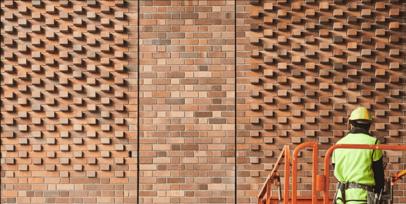 A worker in a yellow helmet and green jacket stands on an orange lift in front of a textured brick wall featuring Lightweight Brick Cladding with a raised, geometric pattern on both sides and flat bricks in the centre. Bricks are Namoi Valley Medium Cream Bricks