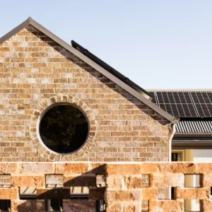 A brick house in Hawthorn 76 Cambridge with a circular window and rooftop solar panels, partially hidden by a brick fence, sits under a clear blue sky.