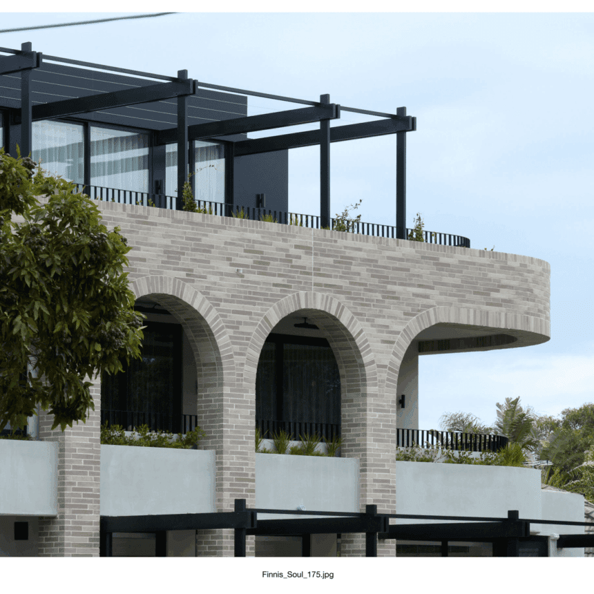 Modern building featuring Bespoke Almond Eco light brick arches on the second floor, large glass windows, and a balcony with black metal railings. Trees and greenery surround the scene beneath a cloudy sky.