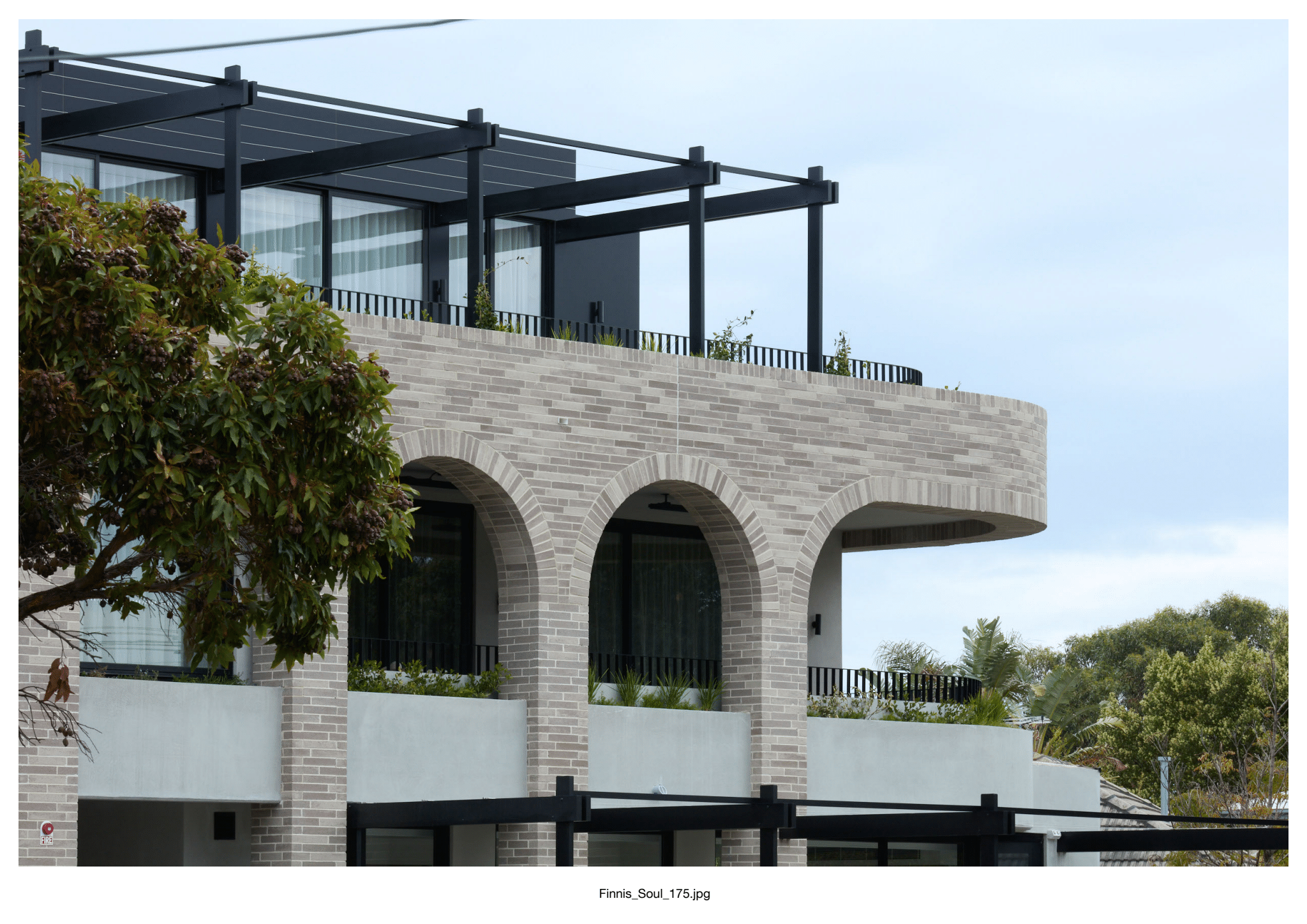 Modern building featuring Bespoke Almond Eco light brick arches on the second floor, large glass windows, and a balcony with black metal railings. Trees and greenery surround the scene beneath a cloudy sky.