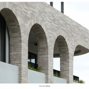 A modern facade with Bespoke Almond Eco light-colored brick arches, black-railed balcony, lush plants, and glass window, set against a bright sky for a refined contemporary look.