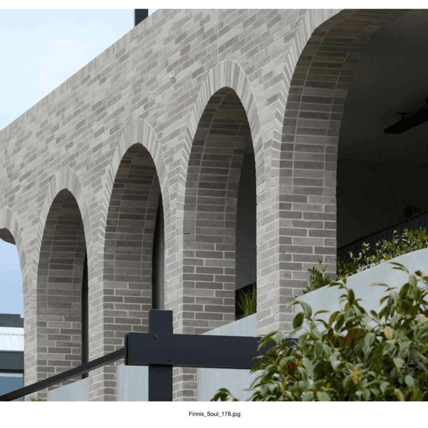 A modern building façade features tall, bespoke Bespoke Almond Eco brick arches, with plants and greenery in the foreground beneath a cloudy sky.