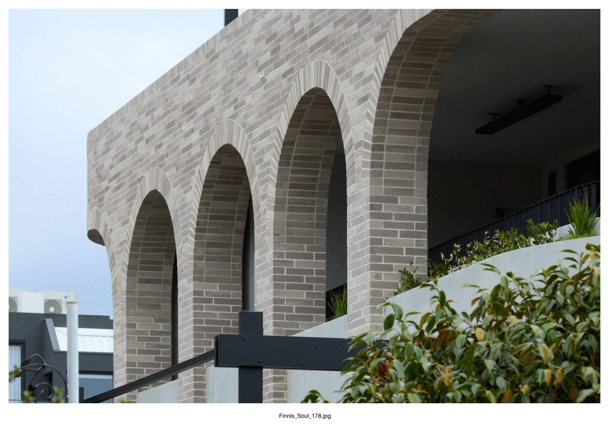 A modern building façade features tall, bespoke Bespoke Almond Eco brick arches, with plants and greenery in the foreground beneath a cloudy sky.
