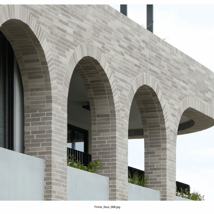 A modern facade with Bespoke Almond Eco light-colored brick arches, black-railed balcony, lush plants, and glass window, set against a bright sky for a refined contemporary look.