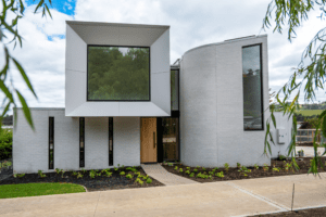Modern two-story house with white brick exterior, large geometric windows, and a curved wall. Breeze block details accent the design. A landscaped garden and path lead to the front door, with trees and greenery in the background.
