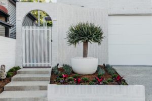 A modern front yard with a white brick wall, breeze block accents, metal gate, and a large potted succulent plant surrounded by mulch and colorful landscaping in front of a white garage.