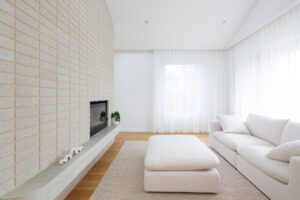 A bright, minimalist living room with a white sofa, matching ottoman, light beige rug, and a modern fireplace set in a breeze block-textured brick wall. Sheer white curtains cover large windows, allowing natural light to fill the space.
