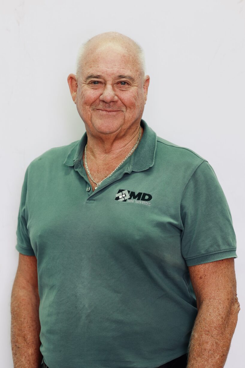 Mick Morley, an older man with a bald head and light skin, wears a green polo shirt with a small MD Brick logo and a silver necklace as he stands smiling against a plain white background.