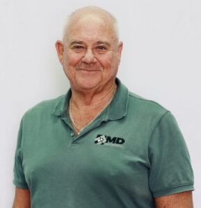 Mick Morley, an older man with a bald head and light skin, wears a green polo shirt with a small MD Brick logo and a silver necklace as he stands smiling against a plain white background.