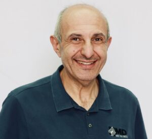 A smiling older man with short gray hair stands against a plain white background, wearing a dark blue polo shirt with an MD Brick logo on the left side.