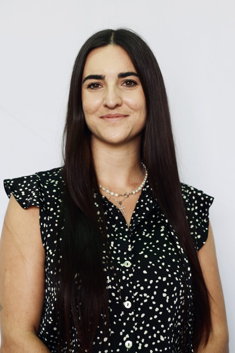 A woman with long straight brown hair, wearing a black blouse with white polka dots and a pearl necklace, stands in front of a plain light background, smiling softly at the camera.