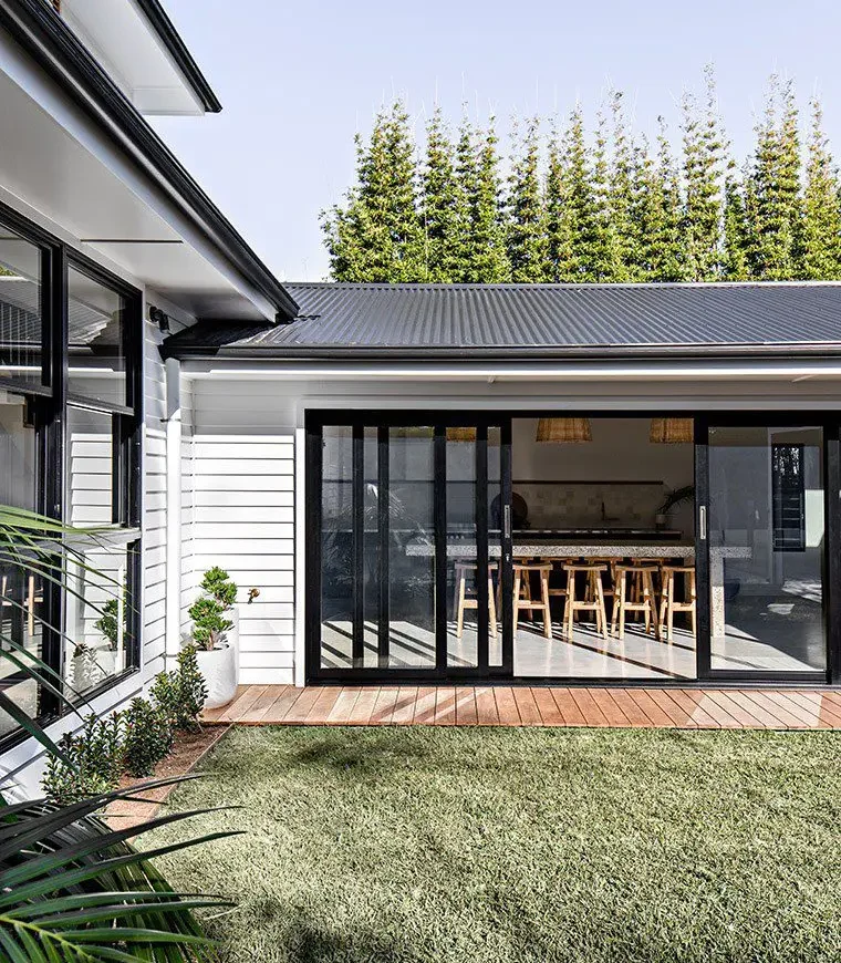 A modern house with white fibre cement siding, large glass sliding doors, a wooden deck, and a grassy backyard. The interior features a wooden dining table with chairs and pendant lights. Tall bamboo plants line the back fence.