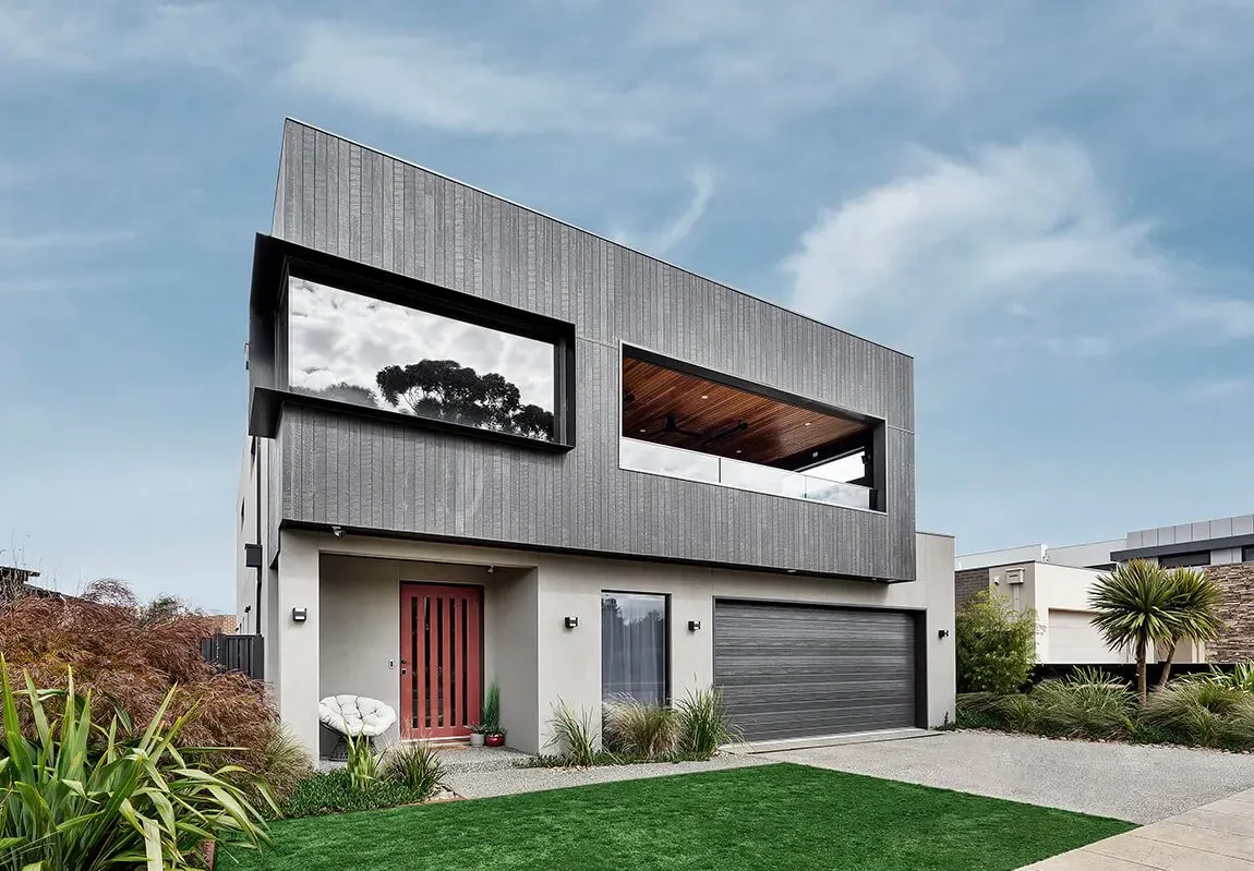 Modern two-story house with a sleek grey fibre cement exterior, large windows, a red front door, double garage, and a landscaped yard with green grass and plants under a partly cloudy sky.