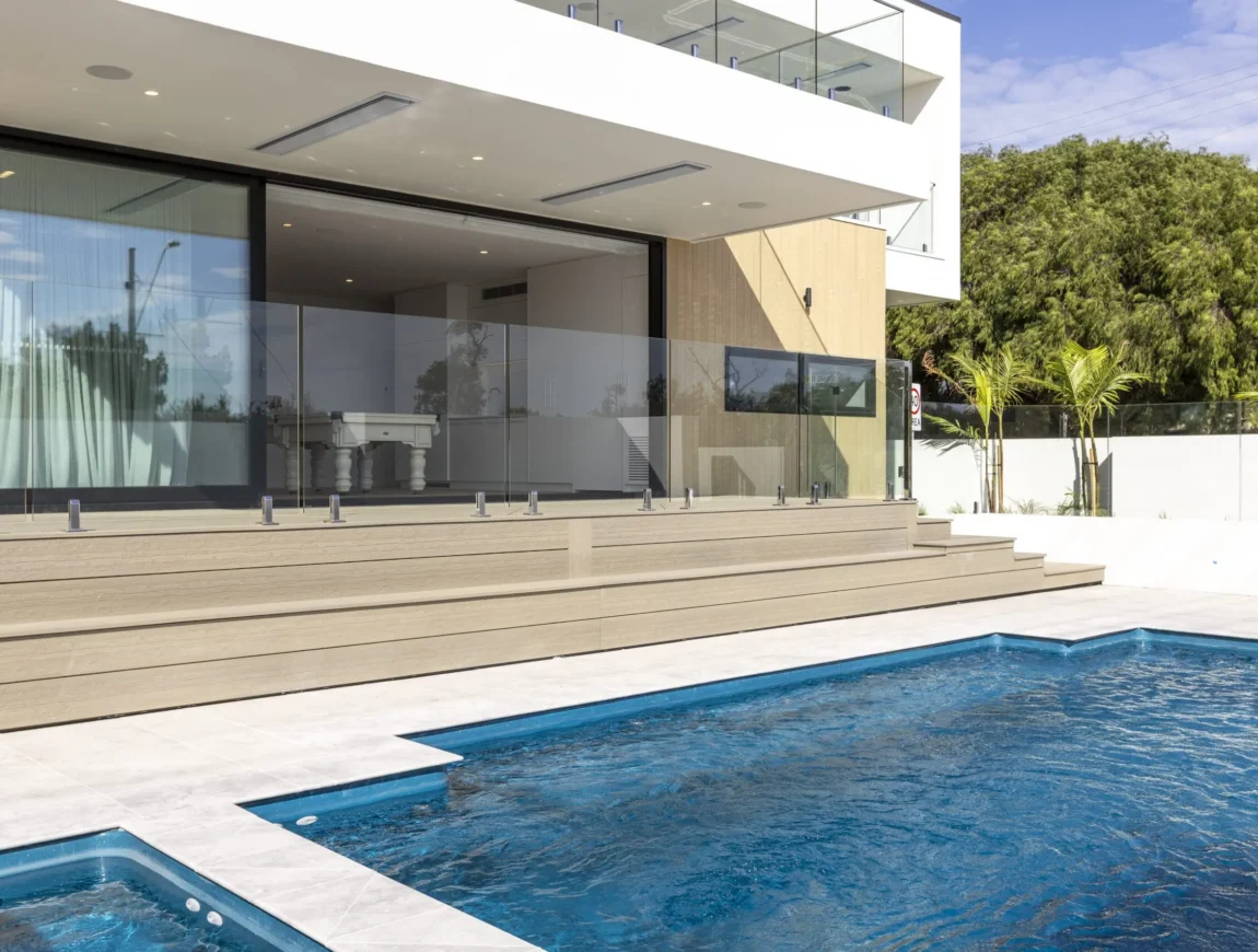 Modern house with large glass doors and balcony, fibre cement accents, light wood steps leading to a patio, and a blue swimming pool in the foreground. Trees and a clear sky are visible in the background.