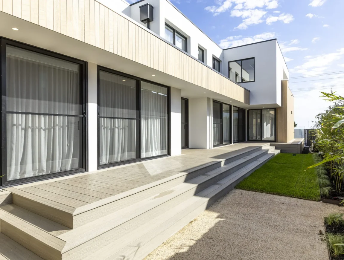 Modern two-story house with large windows and sliding glass doors, light wood paneling and fibre cement accents, plus a spacious deck with wide steps leading to a green lawn and stone path, under a partly cloudy sky.