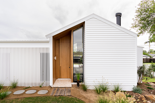 A modern house with white fibre cement siding, a wooden entrance, large vertical window, and minimalist landscaping featuring stepping stones and green plants, all set under a cloudy sky.