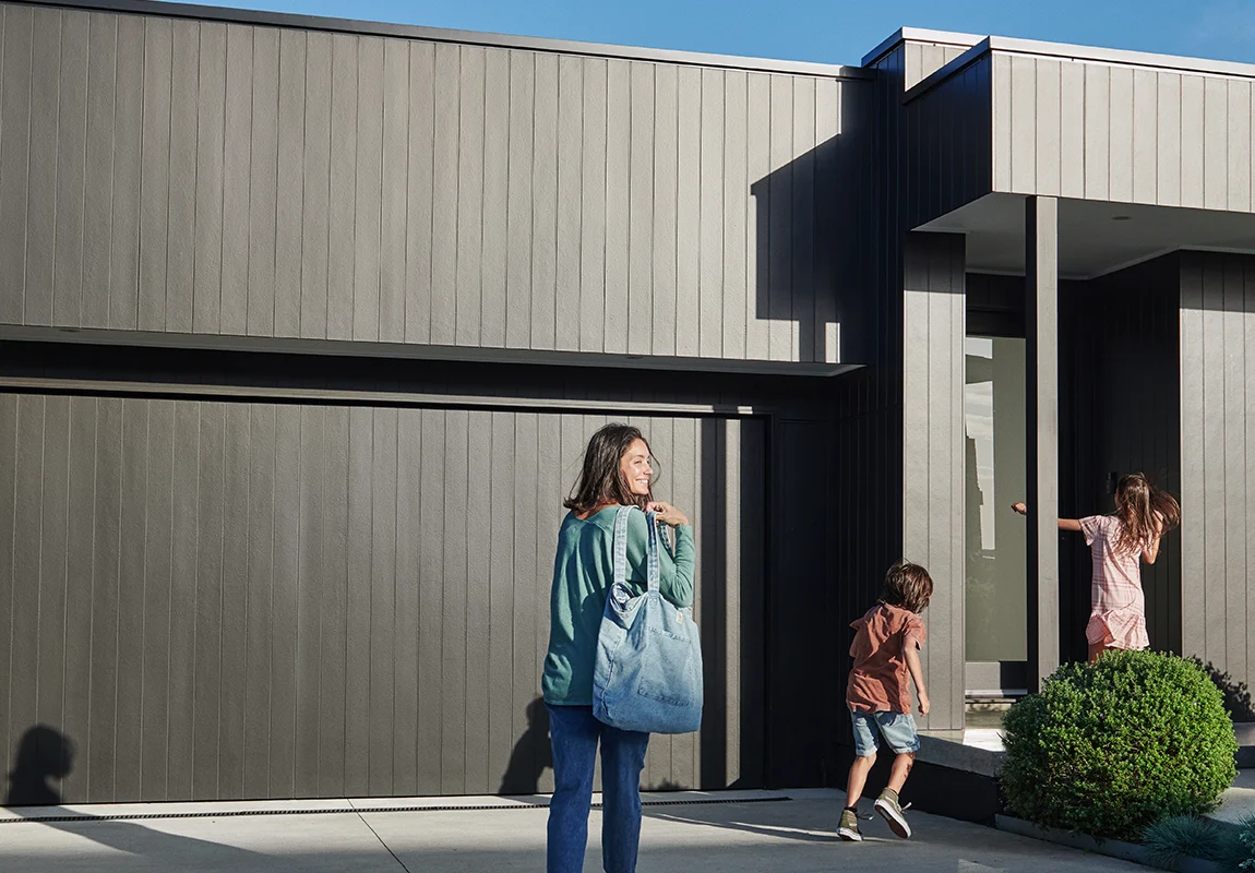 A woman with a bag walks toward a modern black fibre cement house while two children run ahead, one reaching the front door. Shrubs and greenery border the home under a clear blue sky.