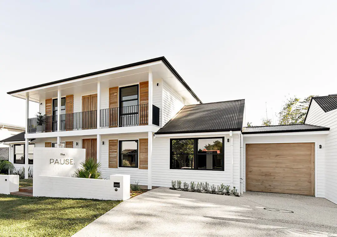 Modern two-story white house with black trim, wood accents, fibre cement siding, a balcony, large windows, and an attached garage. A sign reading The Pause is displayed in the front yard. The sky is clear.