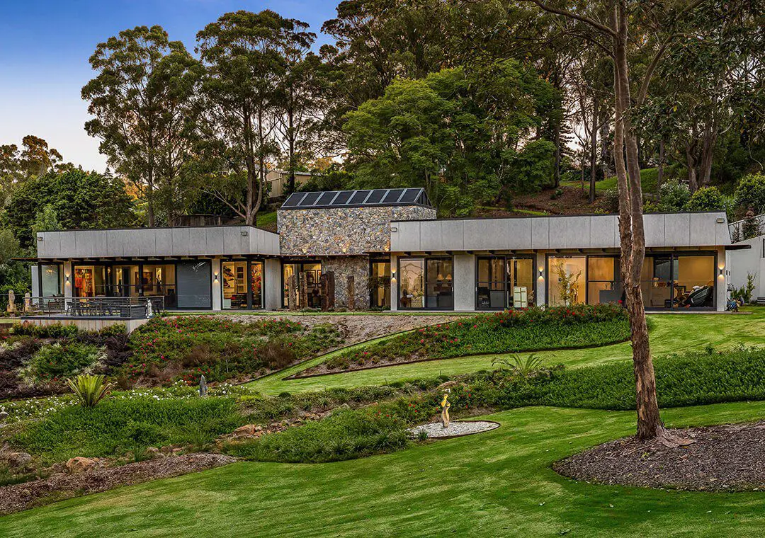 Modern single-story house with large windows, stone accents, and fibre cement siding, surrounded by lush green lawns, landscaped gardens, and tall trees in a serene, wooded setting at dusk.