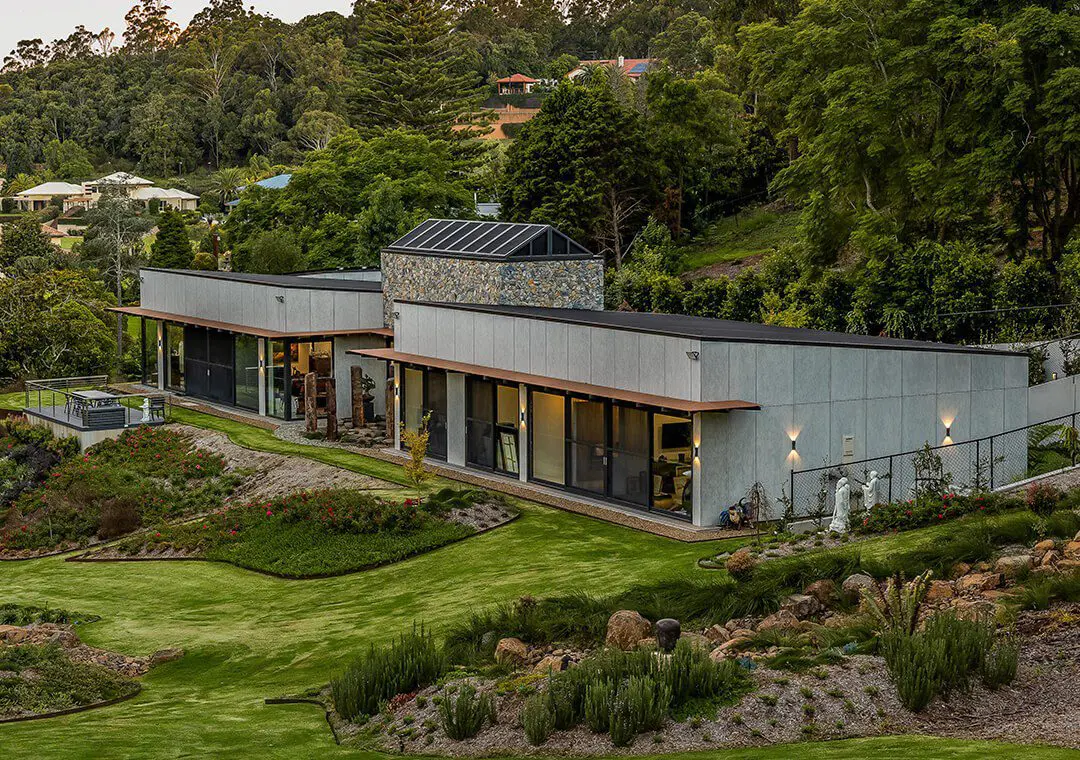 Modern, single-story house with large glass windows and fibre cement and stone accents, surrounded by lush landscaping, green lawn, gardens, and trees in a hilly, residential area.