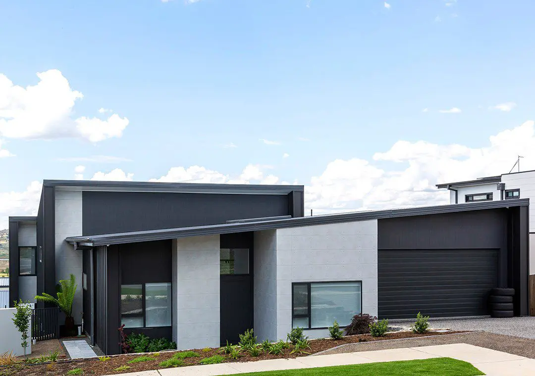 A modern single-story house with a flat sloped roof, black and white fibre cement exterior, large windows, a double garage, and minimal landscaping in the front yard under a partly cloudy sky.