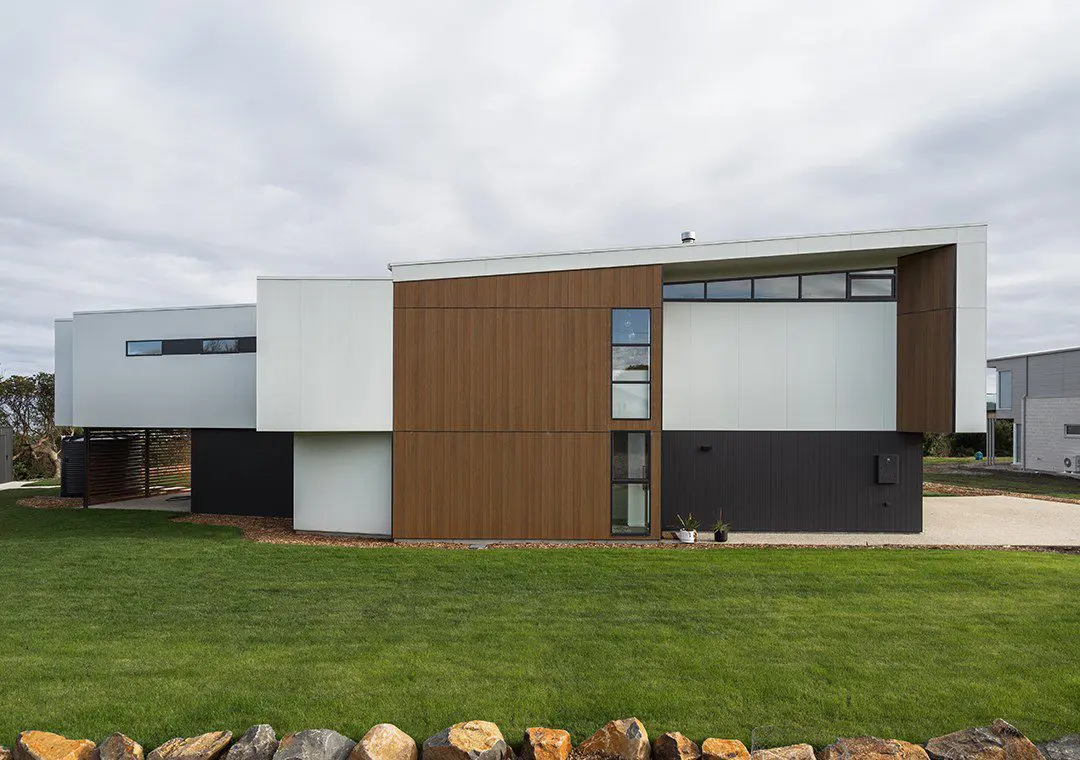 Modern two-story house with a minimalist design, featuring white and wood-paneled fibre cement exterior walls, large windows, and a sloped roof. The house is surrounded by green grass and a stone border.