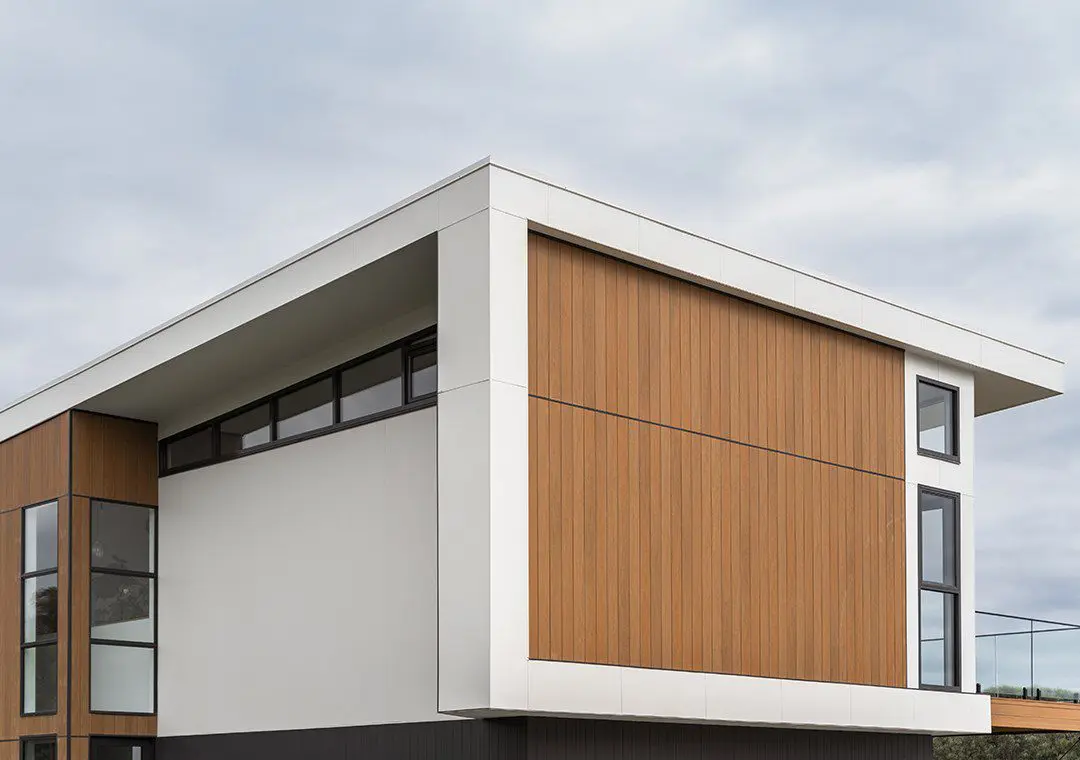 A modern building with clean lines, featuring large windows and a combination of white and wood-textured fibre cement exterior panels under a cloudy sky.