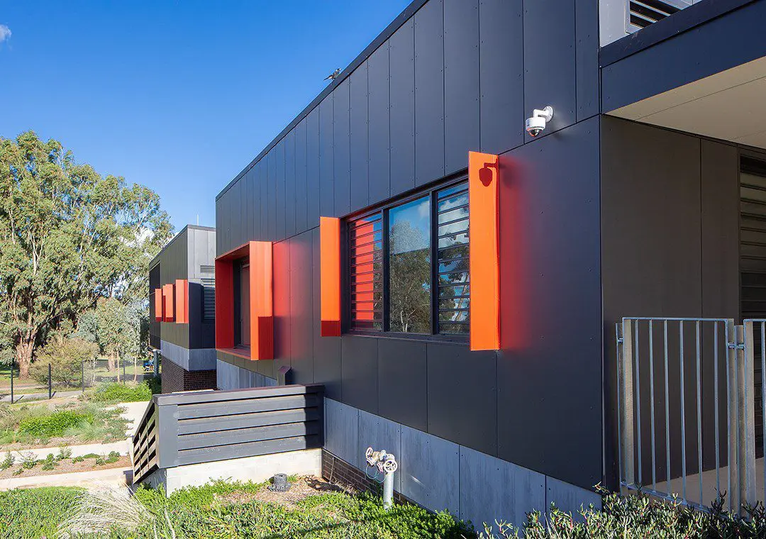 A modern building exterior with dark gray fibre cement paneling and bright orange window accents. There are louvered windows, a metal railing, security camera, and landscaped greenery under a clear blue sky.