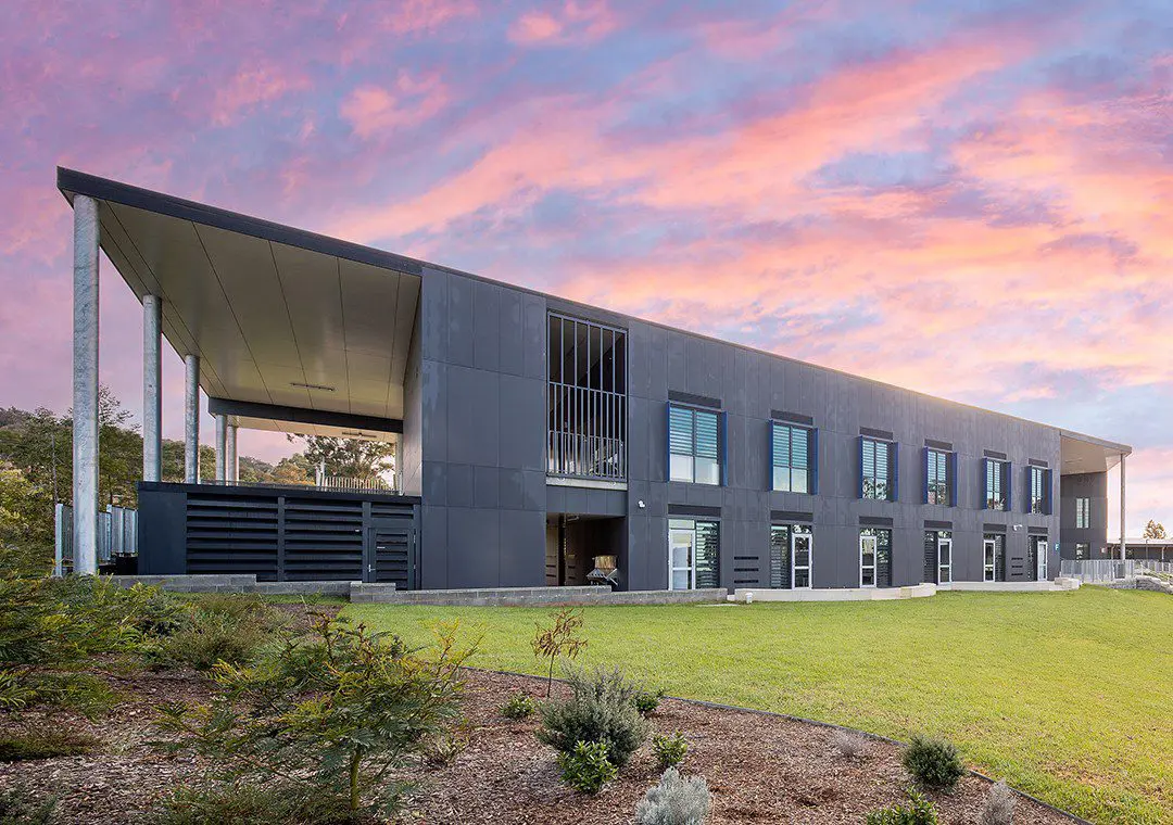 A modern two-story building, featuring sleek fibre cement panels, boasts large windows and a flat, angled roof. It sits on a grassy lawn under a vibrant sunset sky, with bushes and small plants thriving in the foreground.