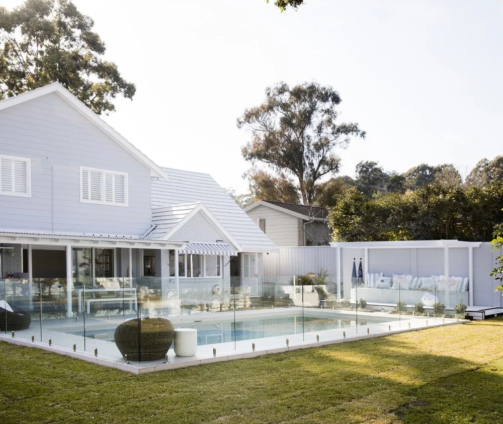 A modern white house with fibre cement accents features a glass-fenced backyard pool, grassy lawn, outdoor seating with cushions, a covered patio, and trees in the background. The scene is bright and sunny.