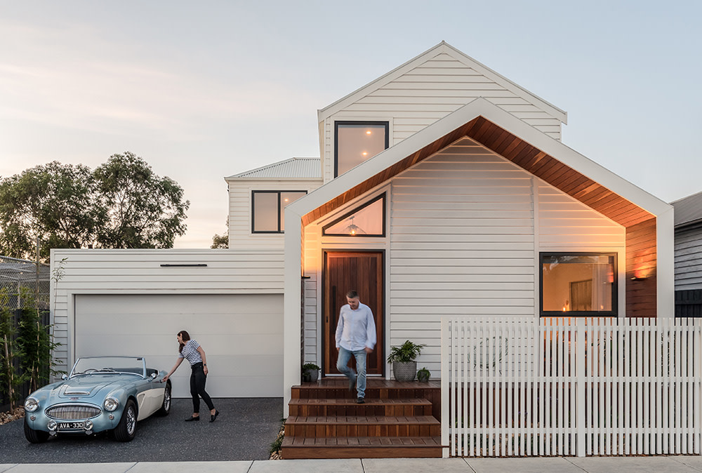 A modern white two-story house with fibre cement and wood accents. A woman stands by a vintage blue car in the driveway, while a man walks down the front steps. The yard is fenced with a classic white picket fence.