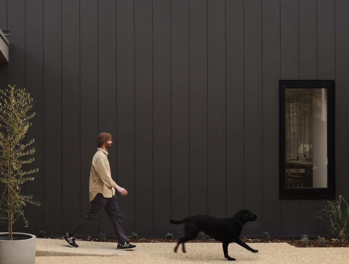 A man in casual clothes walks alongside a black dog on a path next to a modern dark gray building with fibre cement siding, featuring a window, potted plant, and some landscaping.