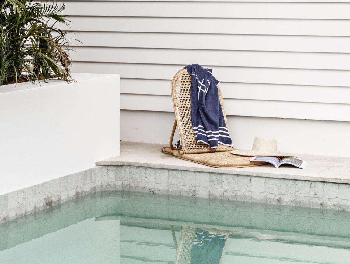 A wicker chair with a blue towel, an open book, and a hat sits beside a clear swimming pool next to white James Hardie Fibre Cement siding and a potted plant.