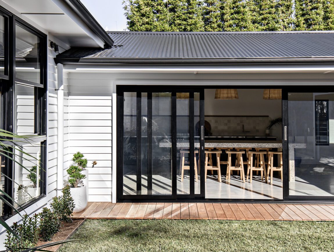 Modern house with James Hardie Fibre Cement white siding and large black-framed sliding glass doors opening to a wooden deck and green lawn; inside, a kitchen island and bar stools are visible. Tall trees line the background.