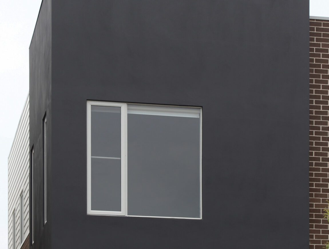 A modern building facade with a large, black rectangular structure featuring a white-framed window. The lower section is brown brick, and part of another section with James Hardie Fibre Cement siding is visible on the left.