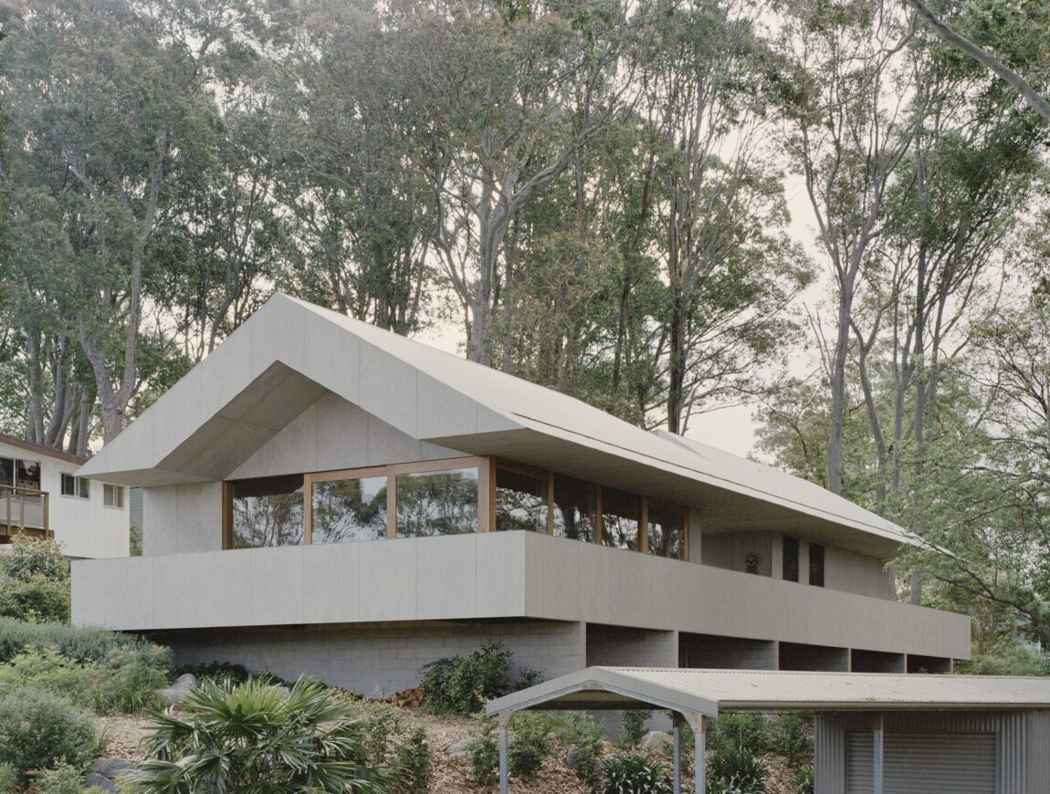 A modern house with a large, angled roof and wide windows sits on a hillside surrounded by tall trees and lush greenery. Durable James Hardie Fibre Cement siding adds to its sleek look. A small carport is visible in the foreground.