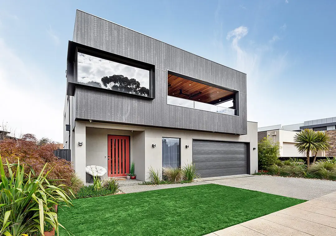 Modern two-story house with gray James Hardie Fibre Cement exterior, large windows, and a double garage. Entry door is reddish-brown with vertical glass panels. The front yard has green grass, plants, and a paved driveway.