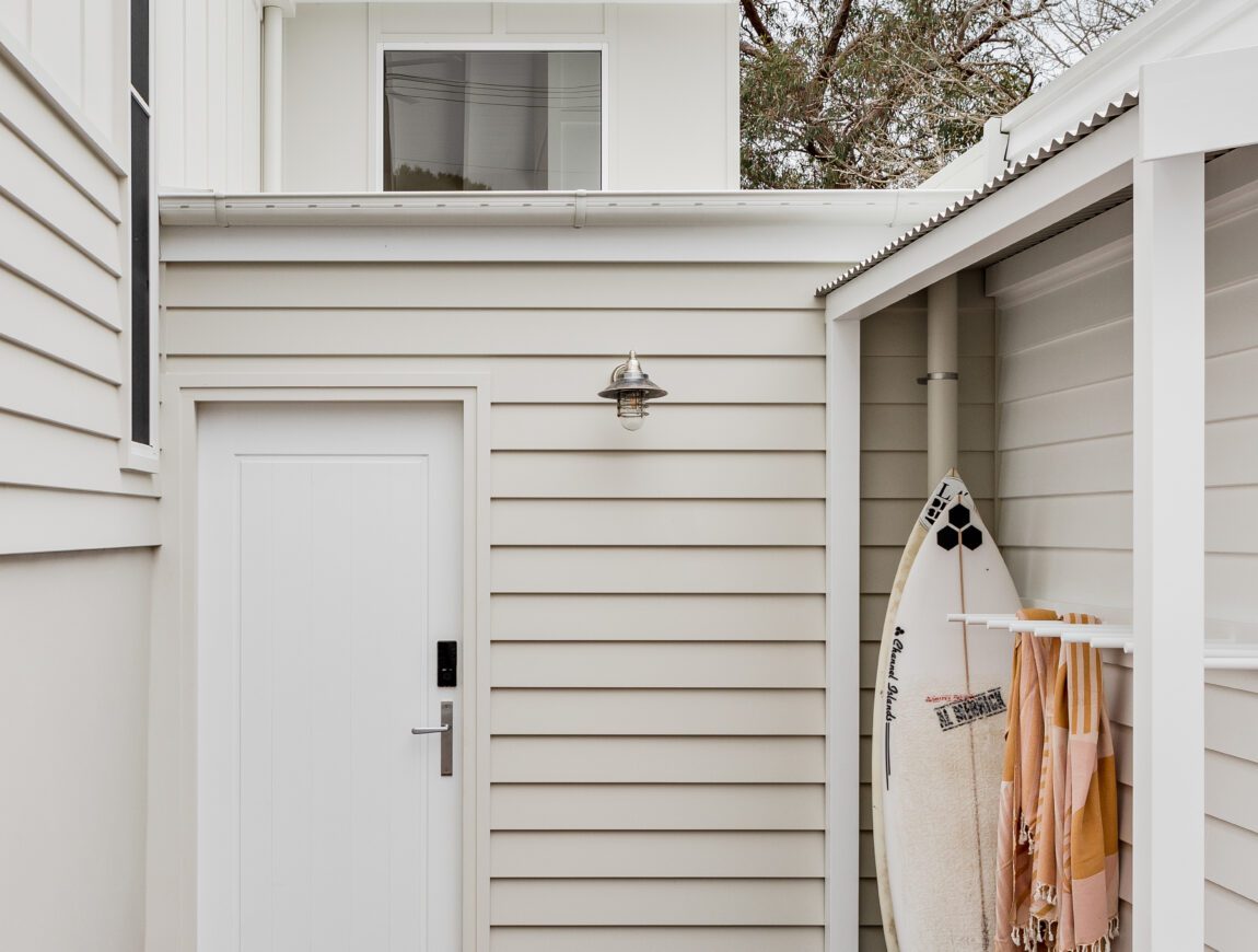 A modern, white-clad exterior made with James Hardie Fibre Cement features a wooden deck, a closed white door, two pairs of sandals, and a surfboard with striped towels beside it. Trees and part of the house are visible in the background.