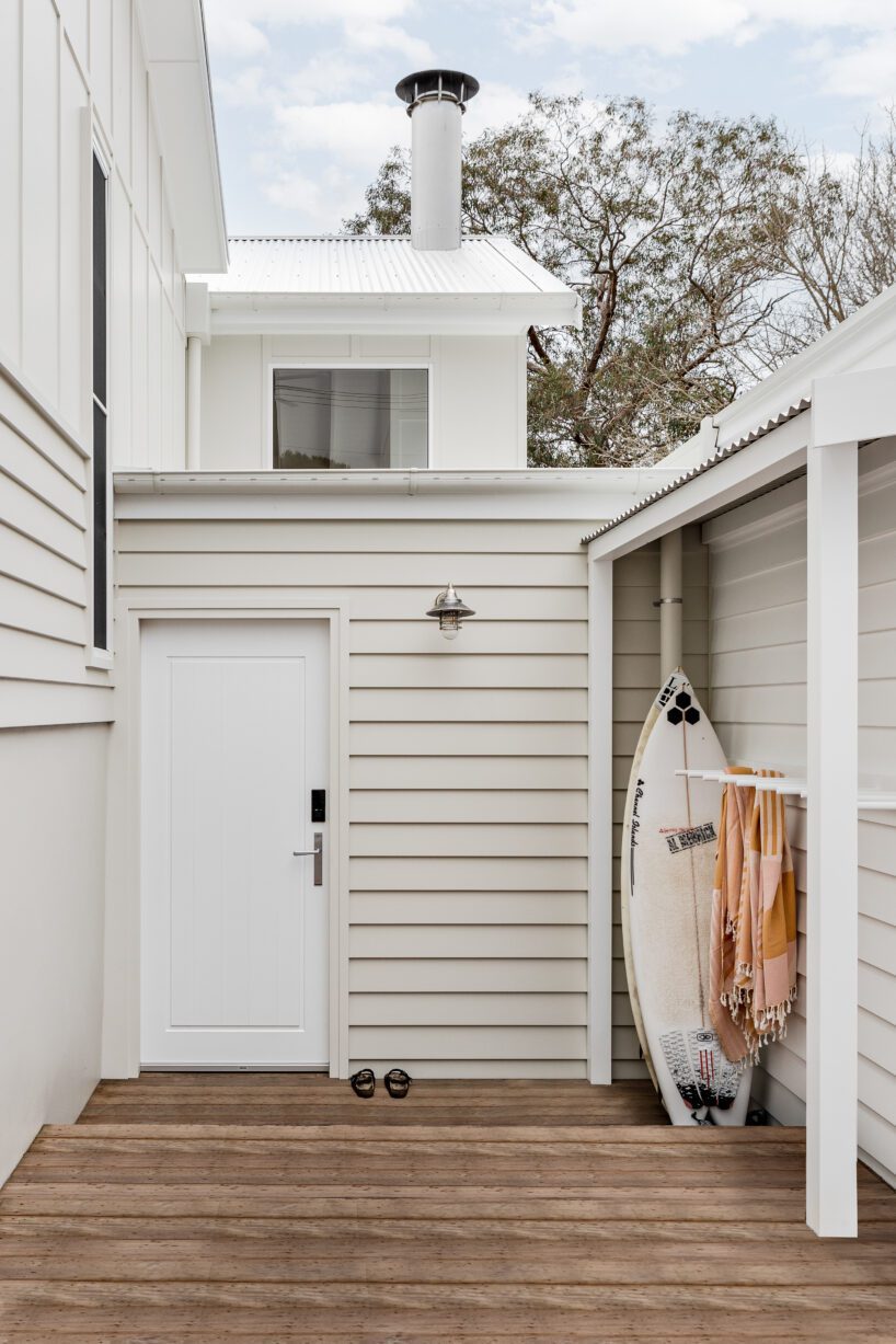 A modern, white-clad exterior made with James Hardie Fibre Cement features a wooden deck, a closed white door, two pairs of sandals, and a surfboard with striped towels beside it. Trees and part of the house are visible in the background.