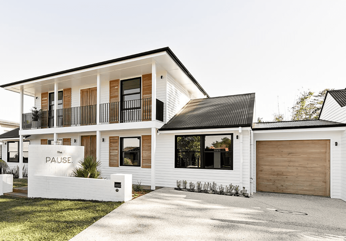 Modern two-story white house with black trim, wooden accents, and durable James Hardie Fibre Cement siding. Features a balcony, single garage with a wooden door, landscaped front yard, and a white PAUSE sign near the entrance.