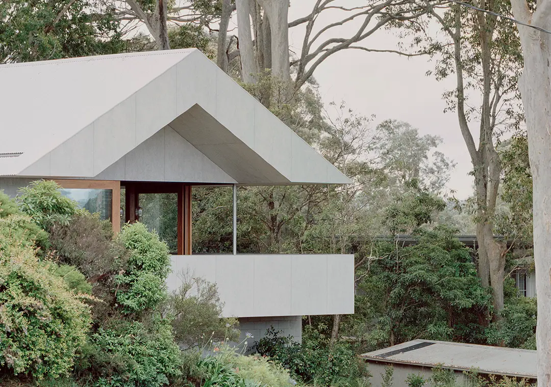 A modern house with a slanted metal roof and a large balcony, featuring James Hardie Fibre Cement siding, surrounded by lush green trees and bushes, blending into a forested environment.