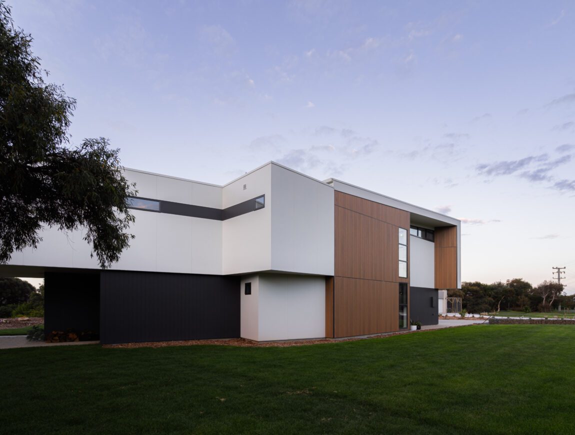 A modern two-story house with a minimalist design, featuring James Hardie Fibre Cement and wood-paneled exterior walls, large windows, and a well-maintained green lawn in the foreground. Trees and a clear sky are in the background.