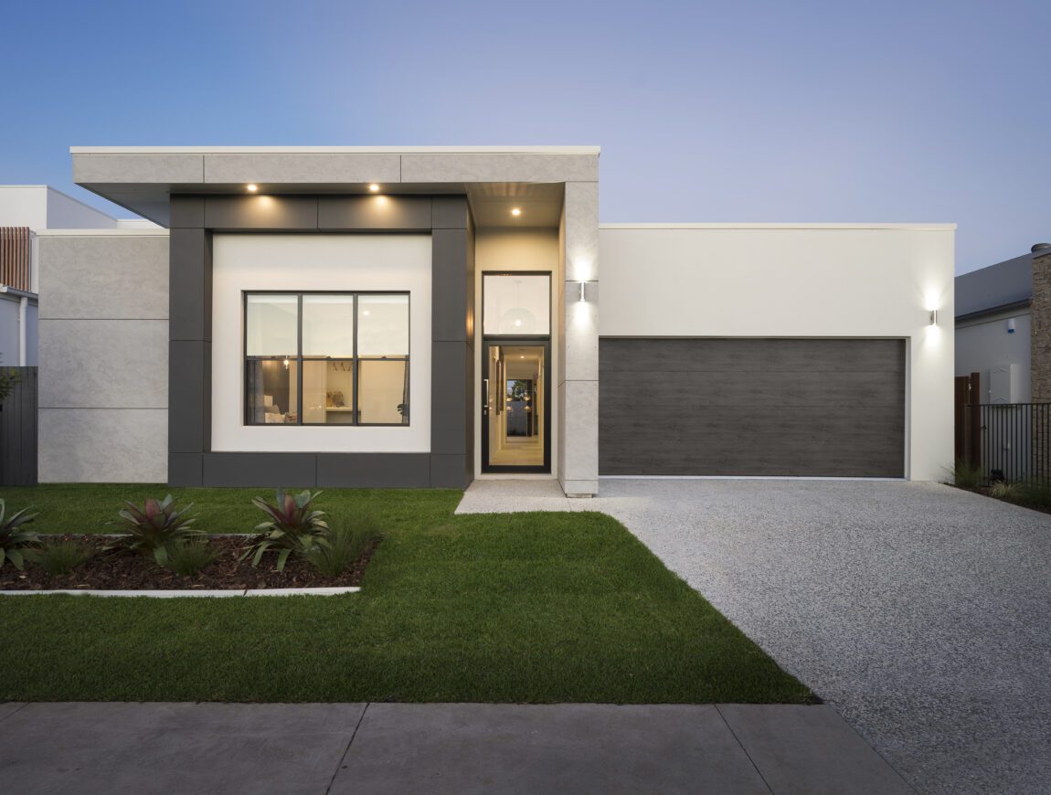 Modern single-story house with large front window, flat roof, and double garage. The exterior showcases clean lines and neutral colors with durable James Hardie Fibre Cement accents, a neatly landscaped yard, and a wide driveway at dusk.