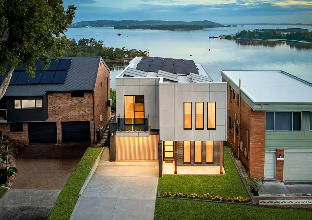 Modern two-story house with large windows and James Hardie Fibre Cement siding, solar panels on the roof, situated between two older homes, facing a calm waterfront with trees and hills in the distance at sunset.