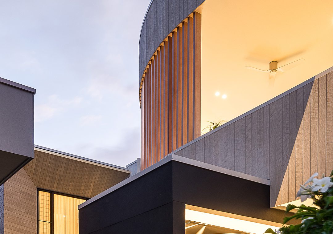 Modern building exterior at dusk with illuminated upper level, curved architectural lines, James Hardie Fibre Cement cladding, wood paneling, and a visible ceiling fan inside. The soft, cloudy sky complements the contemporary design.