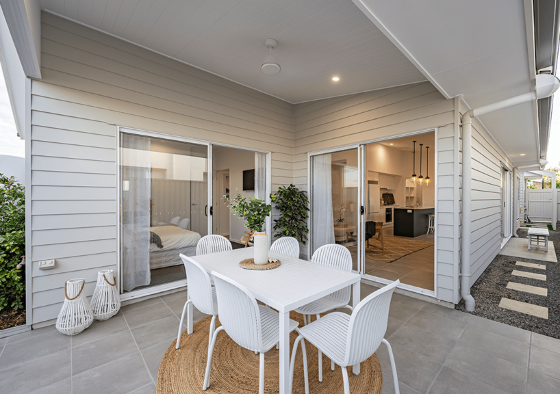 Modern outdoor patio featuring a white dining table and chairs on a round rug, adjacent to a house with James Hardie Fibre Cement siding. Neutral tones and greenery create a cozy, inviting atmosphere connected to the bedroom and kitchen.
