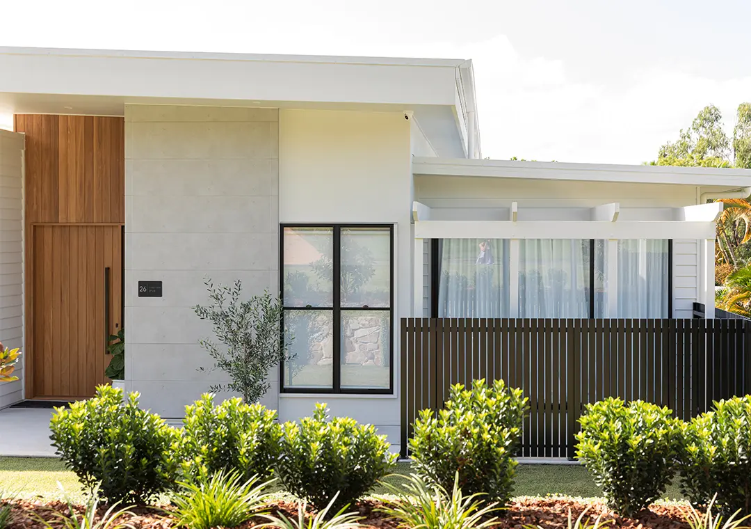 Modern house exterior with light-colored James Hardie Fibre Cement walls, large windows, a wooden door, vertical slat fencing, and neatly landscaped greenery in the front garden.