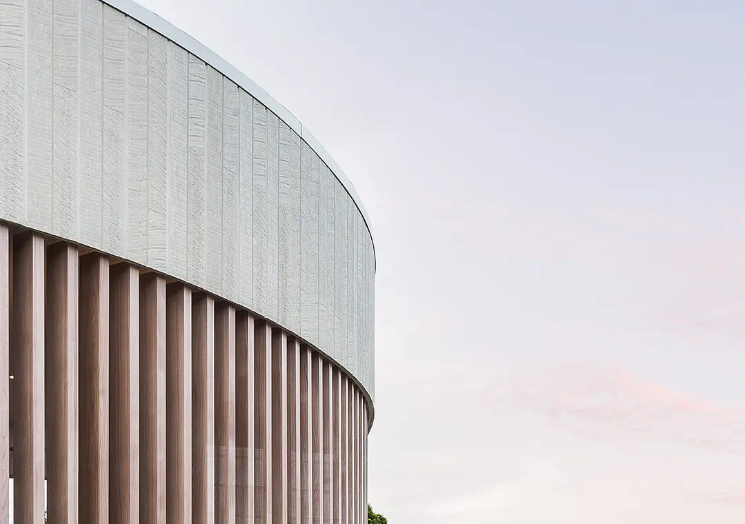 A modern building with vertical wooden slats and a curved white upper facade made from James Hardie Fibre Cement is shown against a pale sky, capturing a minimalist architectural style.