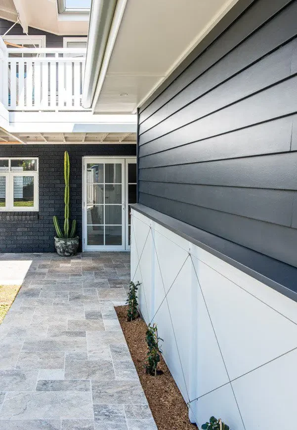 Modern outdoor entryway with gray tiled flooring, James Hardie Fibre Cement dark horizontal siding, white trim, and small plants along the wall. A tall potted cactus is near glass doors, and a white balcony is visible above.